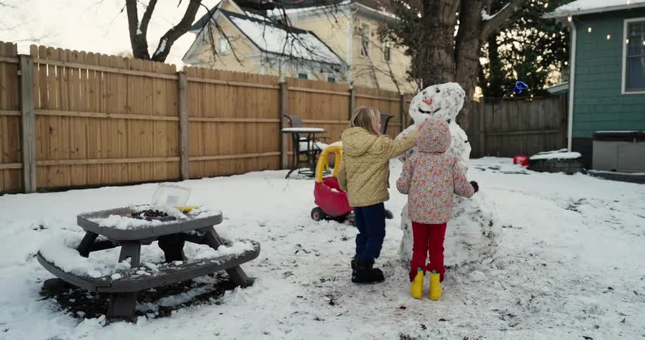 Two young children wearing winter jackets play with a large snowman in a snowy backyard during a winter day