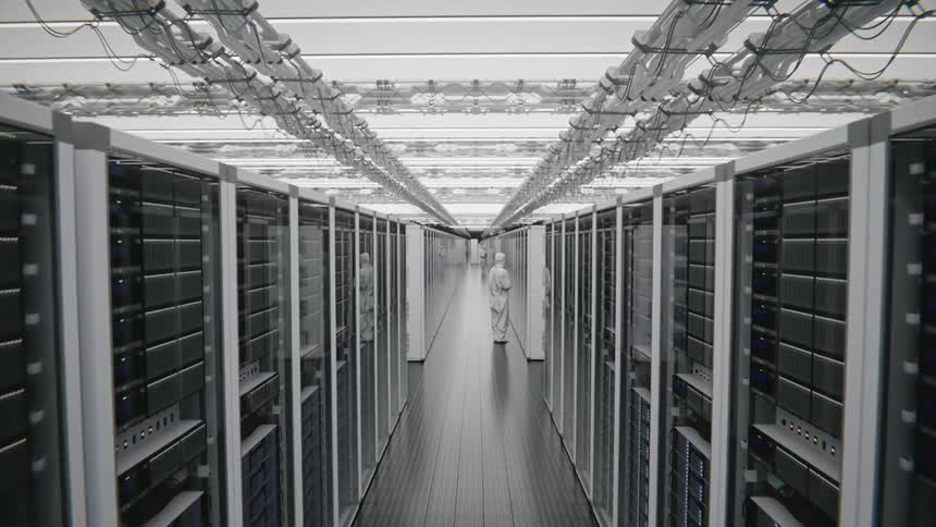 A worker wearing a white protective suit stands between long rows of server racks in a bright data center while the camera pulls back along the central hallway