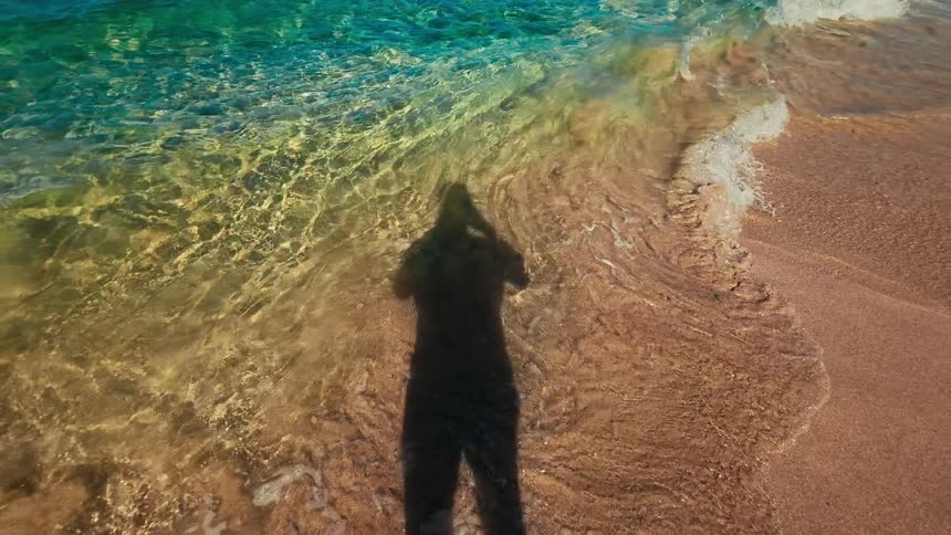 The shadow of a person appears on a sandy beach as clear turquoise water from the ocean ripples over the shore in bright sunlight