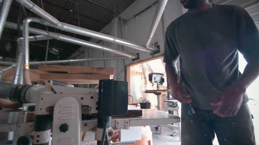 A man wearing a cap and glasses operates a large industrial planer to smooth a thick wooden slab inside a bright workshop filled with metal pipes and various lumber pieces stacked in the background