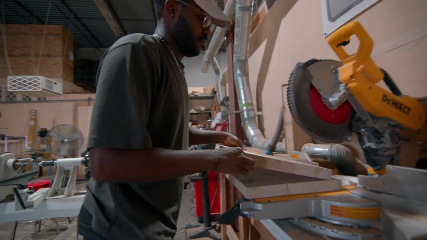 A black man stacks boards near a table saw in a carpentry shop