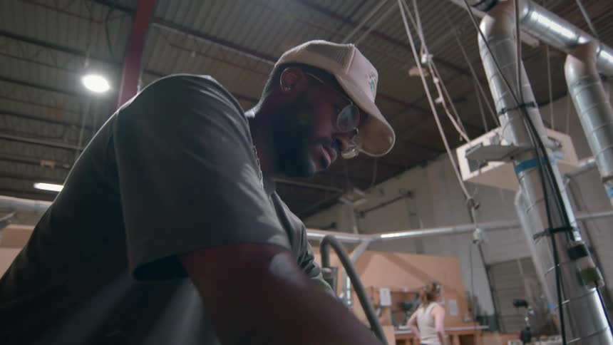 Man standing over a table in a workshop as a woman works in the distance