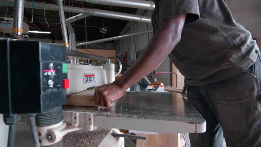 A carpenter in a grey shirt and cap feeds a wooden board through an industrial planer in a woodworking shop