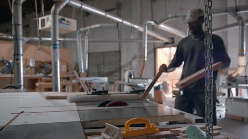 A carpenter wearing a tan cap and glasses works in a professional wood shop as he walks toward a table saw and carefully places two thick boards of light wood onto the work surface