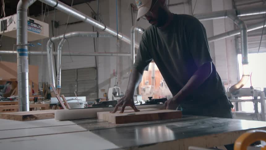 A carpenter wearing a tan cap and glasses works in a workshop as he carefully slides a thick slab of wood across a table saw to make a precise cut