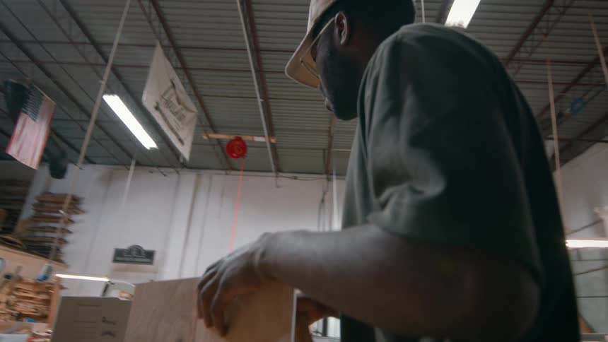 A man working in what appears to be a woodworking shop or warehouse he is seen adjusting a piece of wood and then standing back to examine his progress