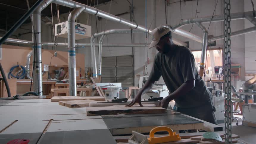 Man operates circular saw cutting wooden board in workshop with overhead ventilation system