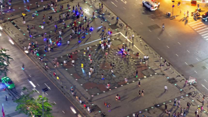 Many people walking across a large patterned stone plaza surrounded by busy city streets with blurred cars passing by while small glowing lights decorate the ground and nearby trees stand under bright street lamps