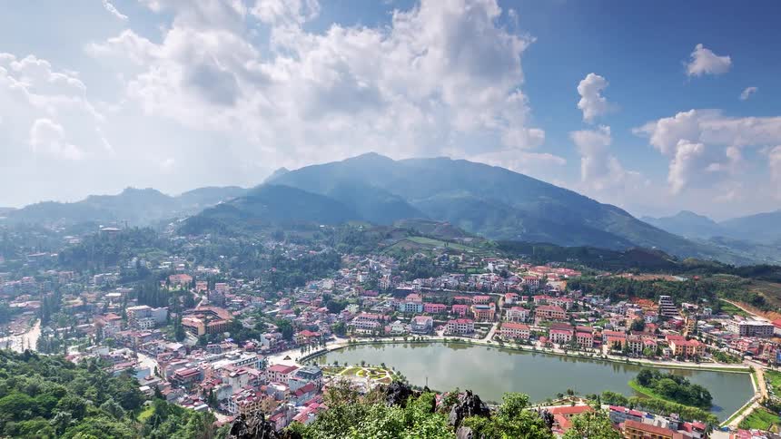 This highangle timelapse captures a scenic town nestled beside a crescentshaped lake with a large misty mountain rising in the background white clouds drift across a blue sky as sunlight shifts over the colorful buildings and lush green hillsides