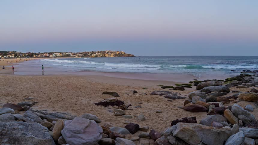 Timelapse captures the transition from a soft pastel sunset to a deep blue twilight over a bustling coastal beachas the city lights begin to flicker on the horizon a bright moon rises and casts a shimmering golden reflection across the ocean waves