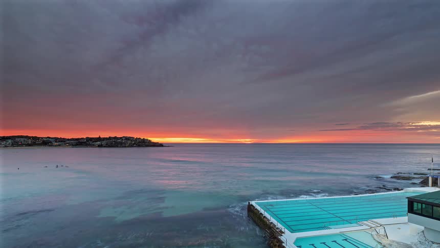 The sun rises over the ocean horizon casting a vibrant red and orange glow across a dramatic cloudfilled skyin the foreground waves crash against the edge of a turquoise oceanfront swimming pool as the morning light gradually brightens the coastal