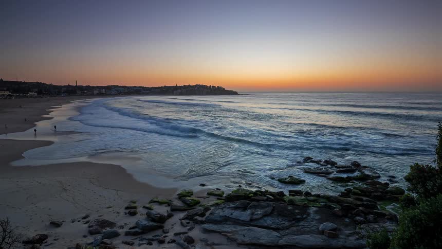 Waves roll in to shore and over the beach as people walk along the coast with neighborhood buildings in the distance as the sun glows on the horizon