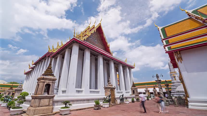 A time lapse view shows a large temple with tall white pillars and an ornate gold and red roof as people walk quickly through the courtyard under a bright blue sky with moving white clouds