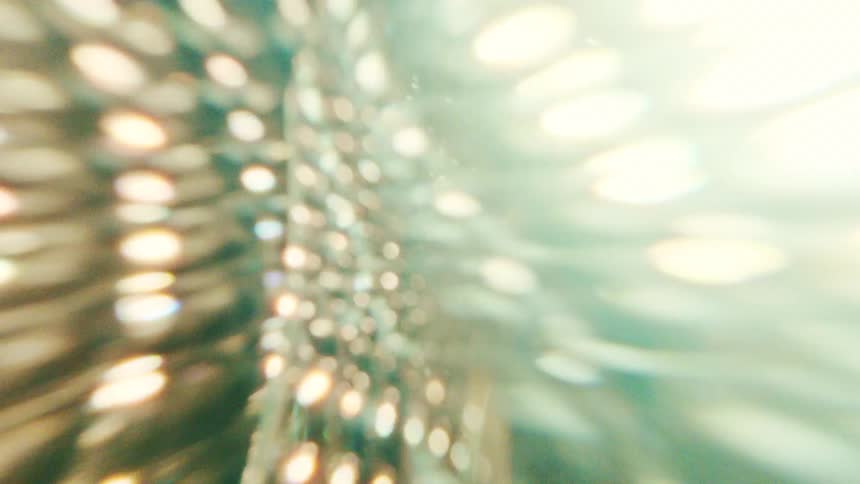A close view shows a pattern of bright circular lights and soft flares that shift and glimmer through a textured glass surface against a dark background creating an abstract glowing effect
