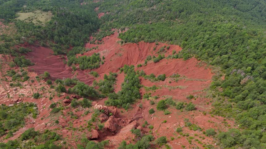 A deep rustcolored canyon cuts through a dense green forest creating a sharp visual contrast between the eroded red earth and the lush canopythe camera pans slowly over the rugged terrain revealing jagged rock formations and steep slopes that highl