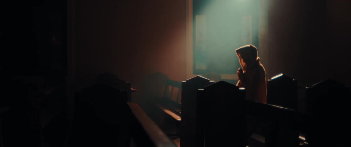 Person wearing a hood is kneeling on a church pew illuminated by a beam of light coming through the window