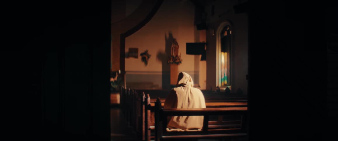 A person wearing a hooded robe sits alone in a wooden pew inside a dark church