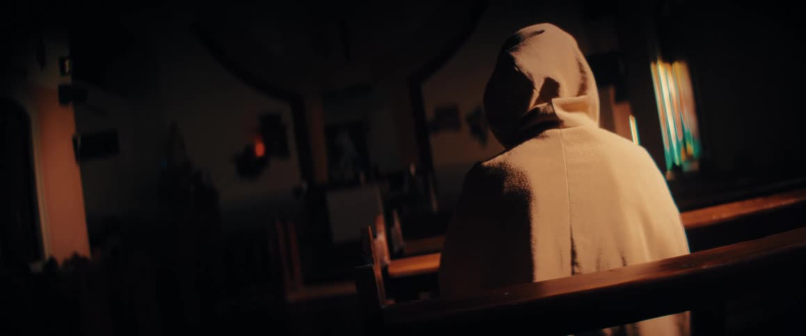 A hooded figure sits in a wooden pew facing away from the camera in a dimly lit atmospheric churchwarm light catches the texture of the persons robe while the background fades into deep shadows and soft architectural details