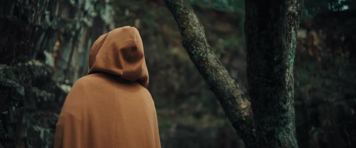 Person wearing a hooded cape stands close to the base of a cliff in the woods on an autumn day