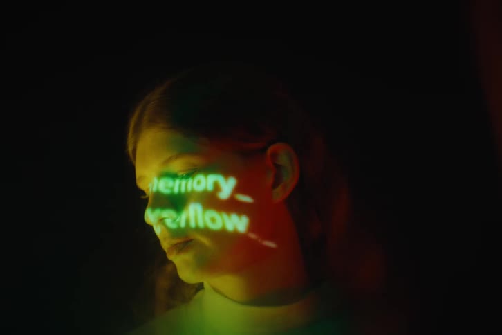 A woman from the neck up is illuminated by warm light as green projected text patterns glow across her face against a dark background