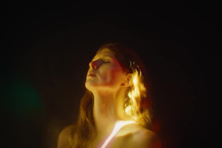 A womans face and upper body are illuminated by shifting abstract light patterns in a dark environment