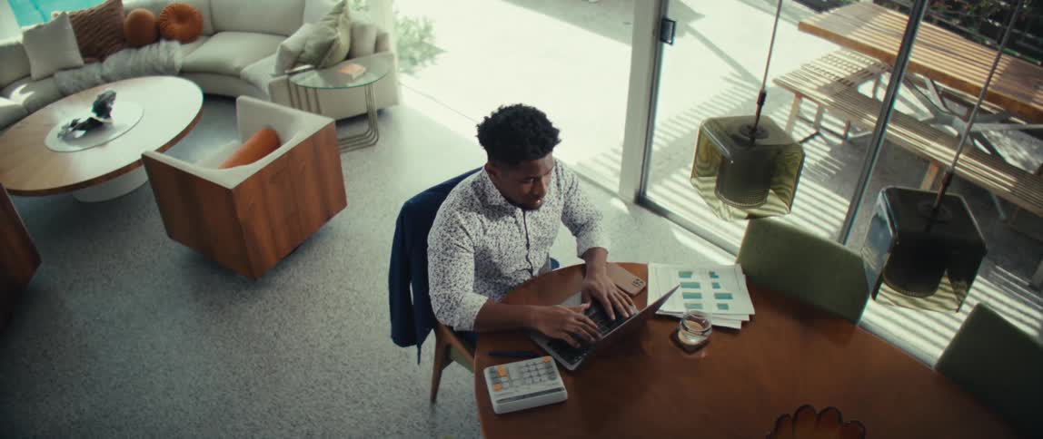 Young adult black man wearing a white dress shirt sitting down and typing on a laptop computer while talking over a wooden table in a living room indoors during day time