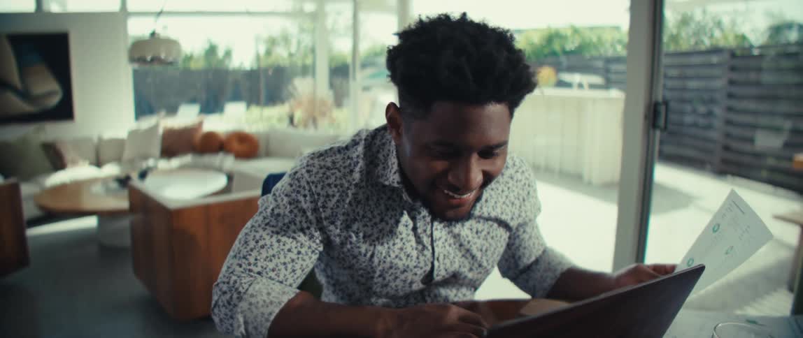 A man smiles while using his laptop and reviewing a document