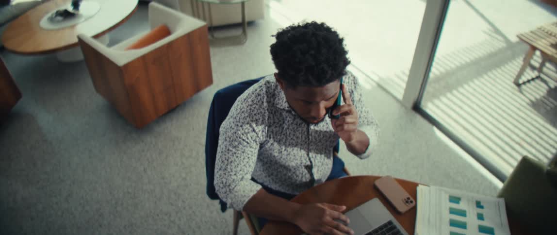 A man talks on a smartphone while using a laptop at a desk