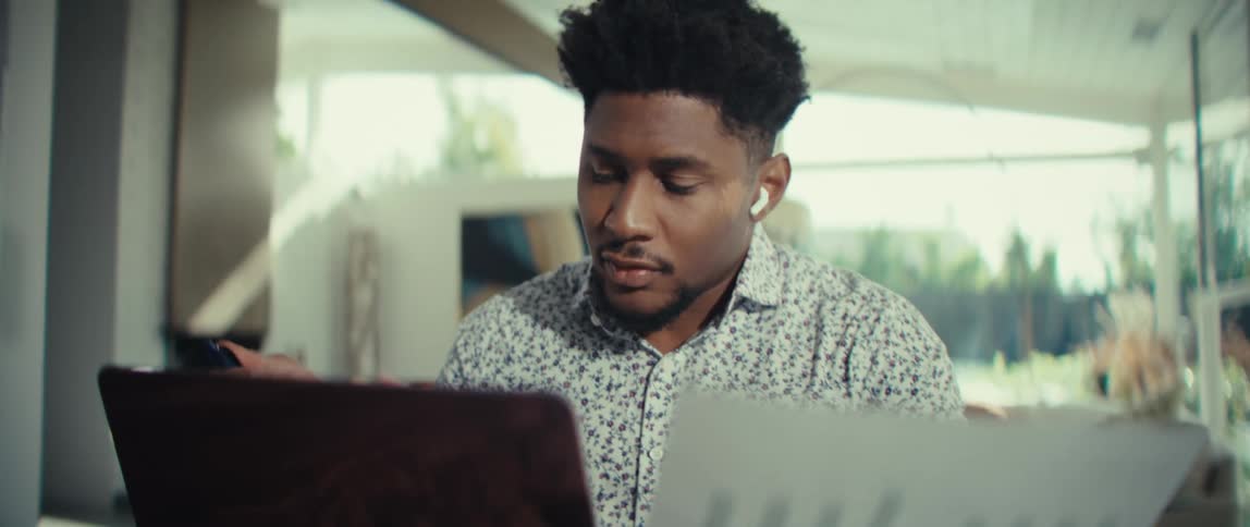 A young black man wearing a floral shirt and wireless earbuds sits at a wooden desk in a bright room while looking closely at a printed bar chart and his laptop screen