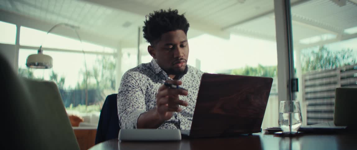 Man sitting at a table in front of a laptop computer playing with his pen as he thinks before typing