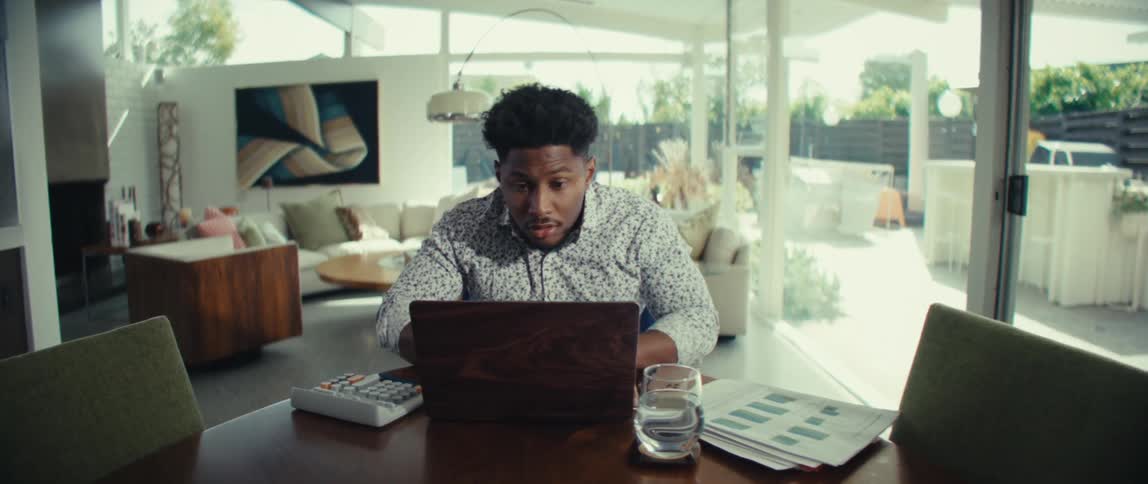 Man works on laptop at table with calculator glass of water and reports in a room with large windows