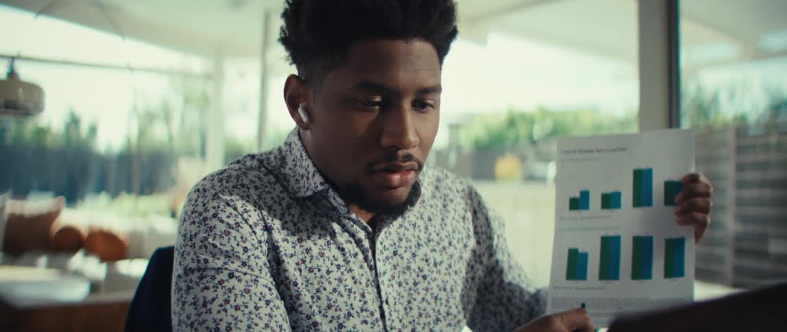 A man in a patterned shirt sits at a dining table presenting a bar chart to his laptop during a video call he uses a pen to point at specific data points on the paper while wearing wireless earbuds to communicate