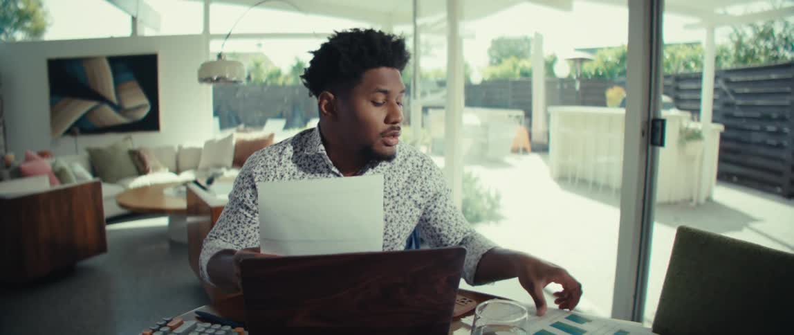 A man wearing a patterned shirt and wireless earbuds sits at a dining table and reads documents with colorful charts during a video call on a laptop in a bright modern home