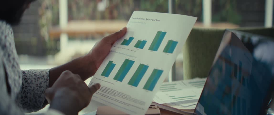 A man holds a page and reviews graphs while seated with his laptop