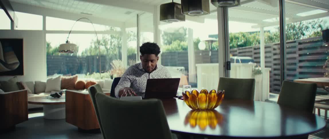 A man in a patterned shirt sits at a dining table looking at his laptop with a mix of confusion and frustration he gestures emphatically with his hands toward the screen while talking appearing to be in the middle of a stressful remote meeting or troub