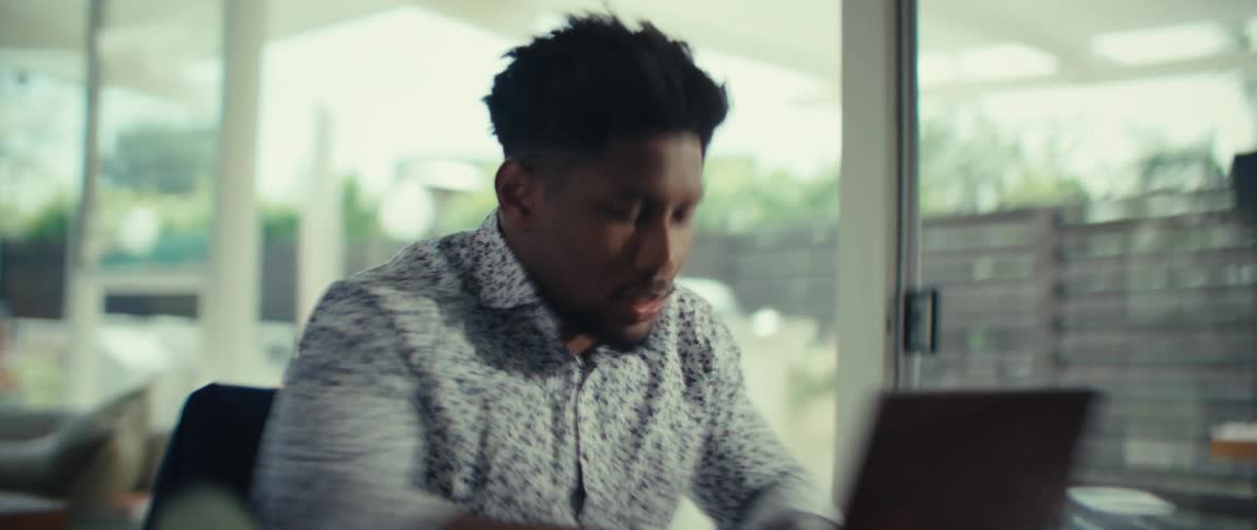 A man with dark hair and a patterned shirt sits at a table in a bright room and reacts with excitement while looking at a laptop computer