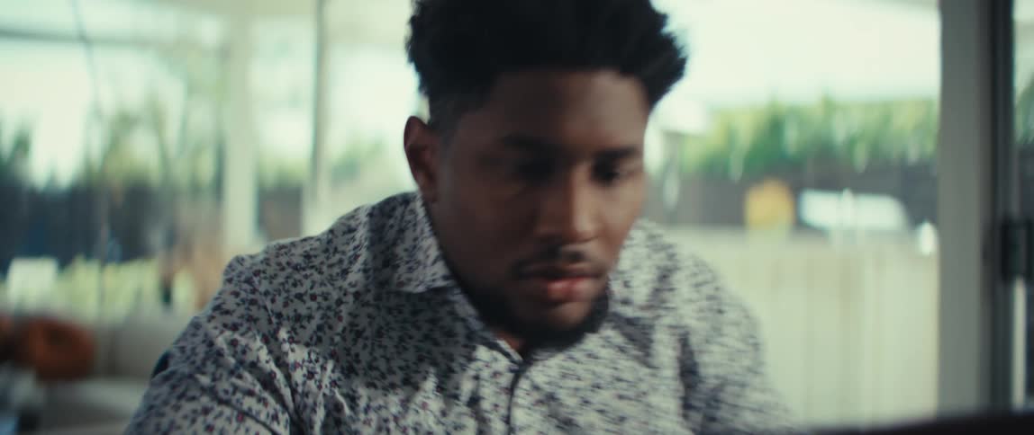 A man sits at a table outdoors focused on working on his laptop as the camera zooms in closer to his face he transitions from a serious expression to a bright satisfied smile