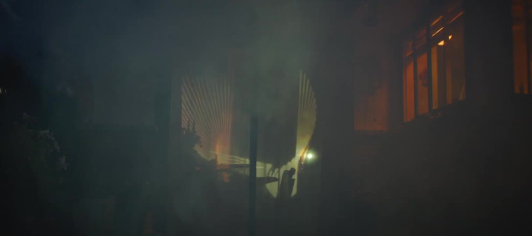 Thick white smoke drifts across the backyard of a house at night as a patterned light projection shines onto a glass sliding door and white curtains