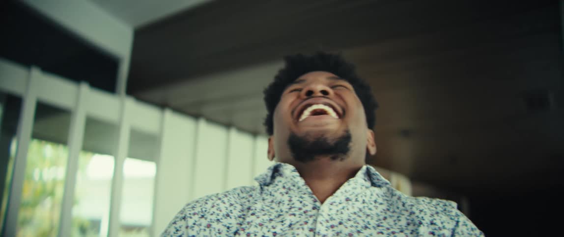 A man wearing a floral patterned shirt looks at his phone and laughs with a wide smile before tilting his head back in a celebratory gesture in a bright indoor setting