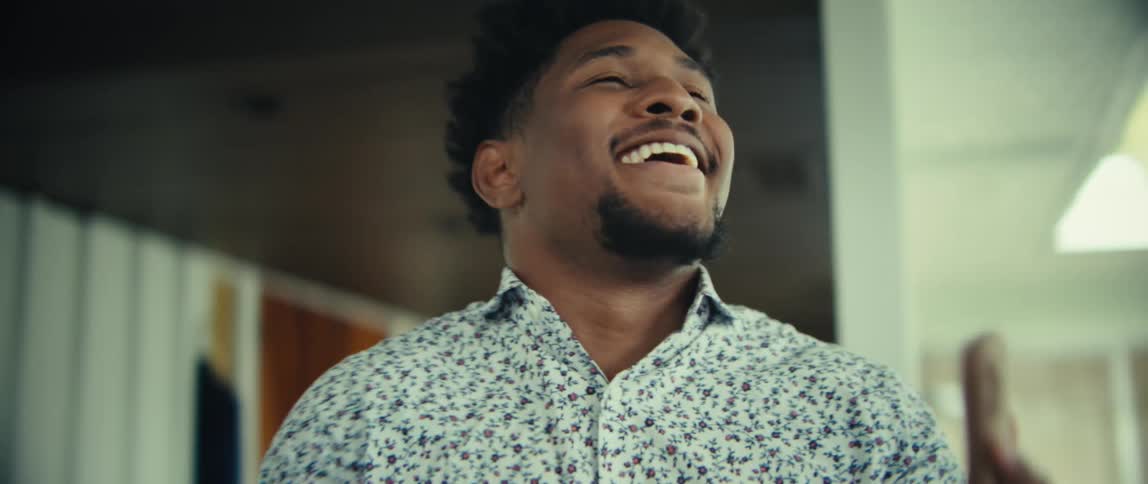 A man wearing a patterned shirt and blue trousers walks through a bright modern house while looking at a phone and laughing with an excited expression