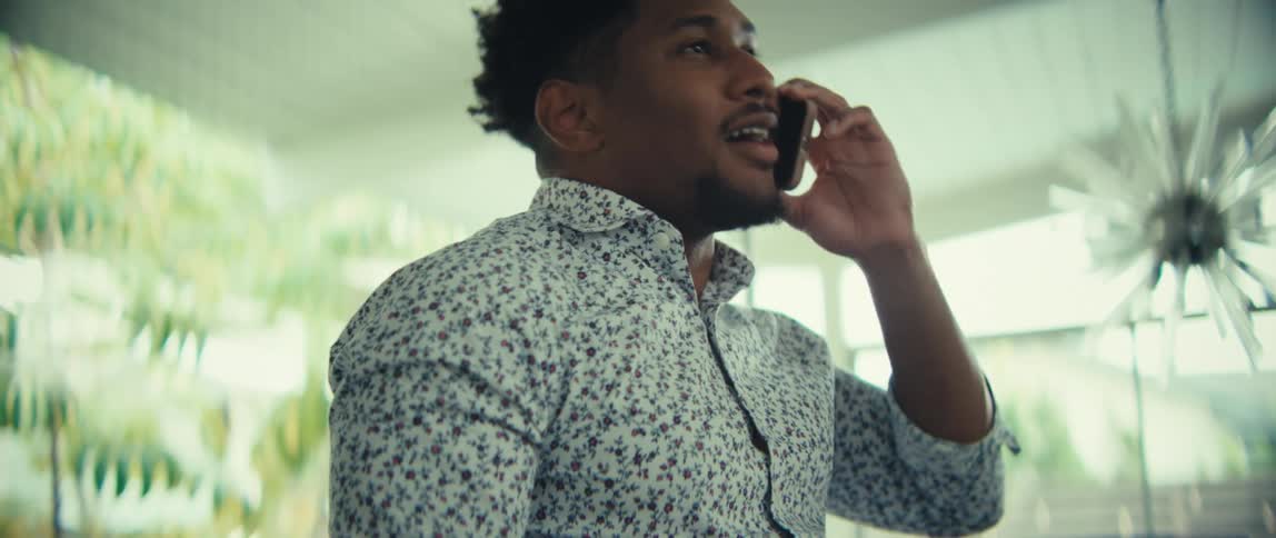 A man wearing a floral shirt speaks on a mobile phone and laughs with a wide smile before pumping his fist in a celebratory gesture while walking through a bright indoor hallway