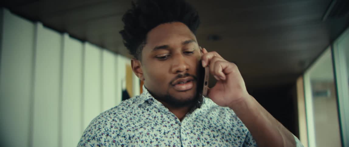 A man wearing a floral shirt and blue trousers speaks on a mobile phone and laughs with a wide smile before hanging a dark suit jacket on a wall hook in a bright modern home
