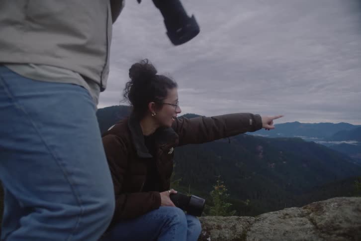 Two tourists stand on a mountaintop at midday both holding digital cameras as a young woman sits on a rock pointing into the distance to the man
