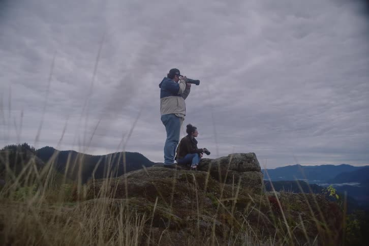 A man standing and a woman sitting on a large rock use cameras to take photos of a mountain range under a cloudy sky during a cold day