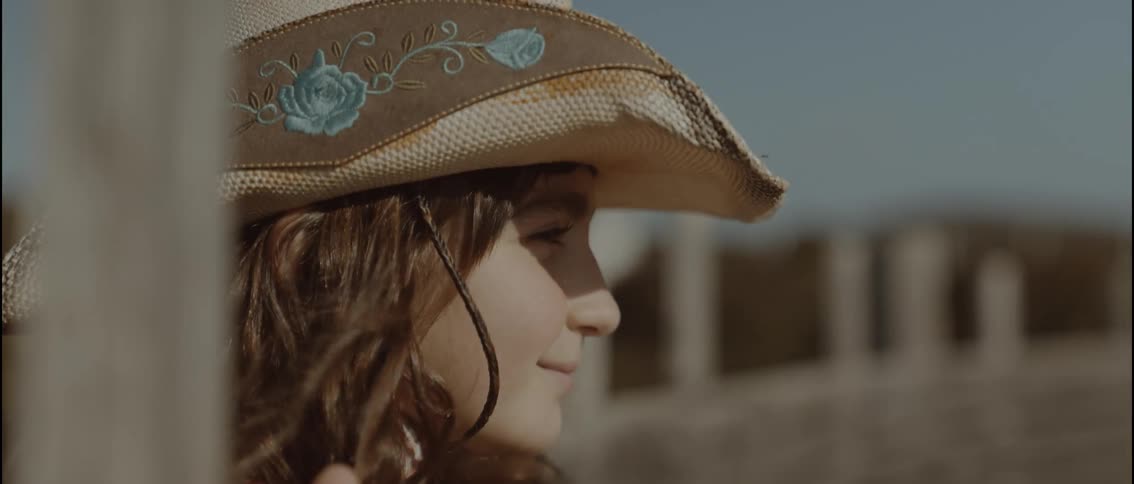 A young girl wearing a brown cowboy hat with blue flowers smiles while looking off to the side in a sunny outdoor setting with a wooden fence and a blurred background