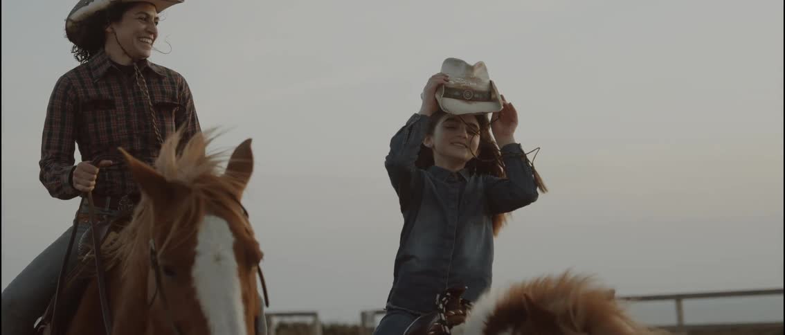 A woman and a young girl laugh while riding horses on a ranch at sunset