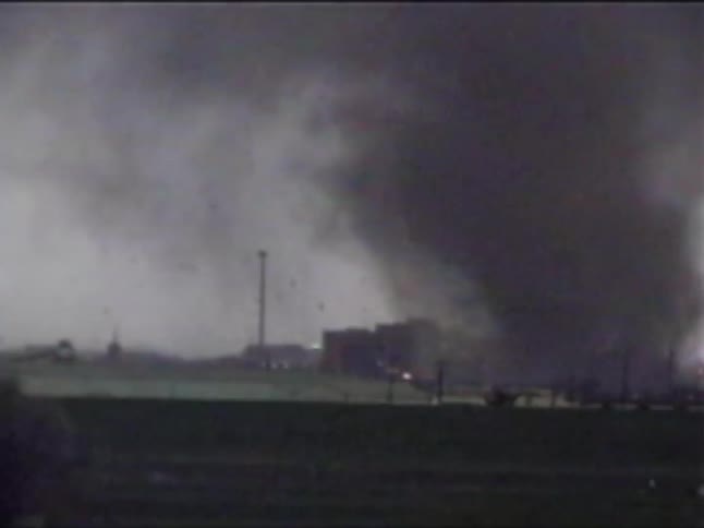 A large dark tornado moves across a landscape kicking up a significant amount of debris as it passes near buildingsthe funnel cloud appears dense and turbulent obscuring the background as it tracks behind a recycling center and toward a