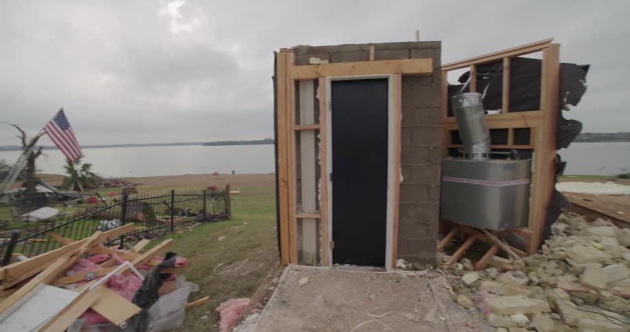 A tracking shot moves across piles of shattered timber and pink insulation to reveal a concrete storm shelter standing solitary and intact amidst the wreckage of a destroyed home