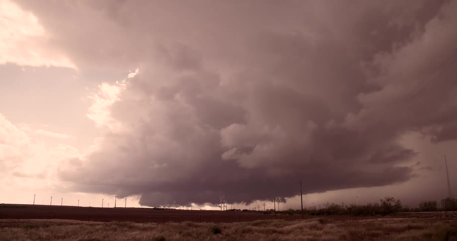 A massive sepiatoned supercell churns violently in a timelapse its rotating cloud structure swirling ominously above a row of tiny wind turbines on the horizon