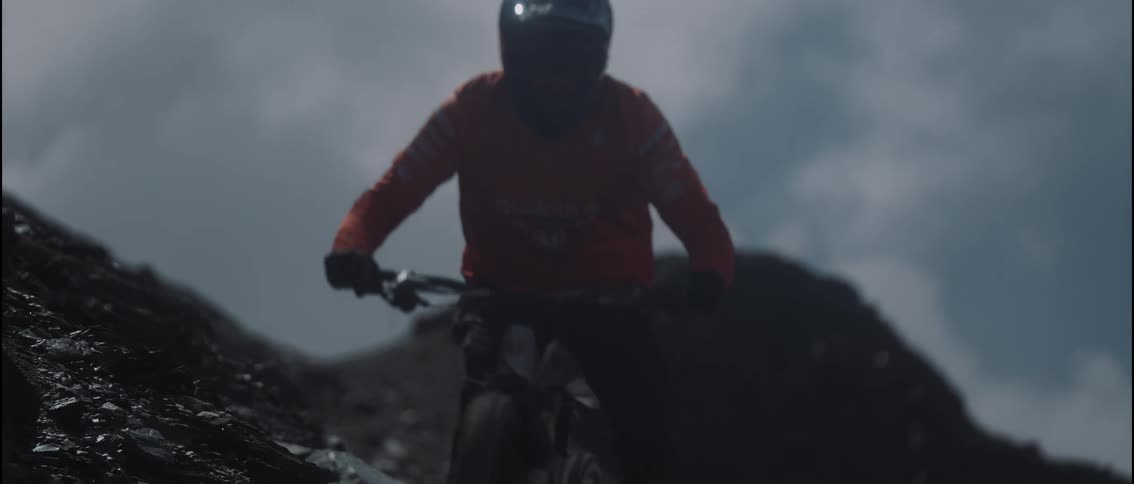 A mountain biker in an orange jersey and fullface helmet descends a steep rocky ridge the scene captures the intense movement as they ride toward the camera against a moody overcast sky